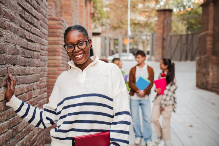 On foreground a african american female student smiling and looking at camera with perfect white teeth standing at university campus. Schoolgirl staring front holding a notebook